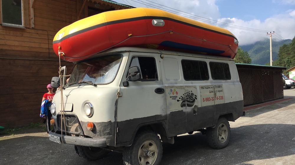 A rubber whitewater raft strapped to the top of a gray Soviet-style UAZ van.  